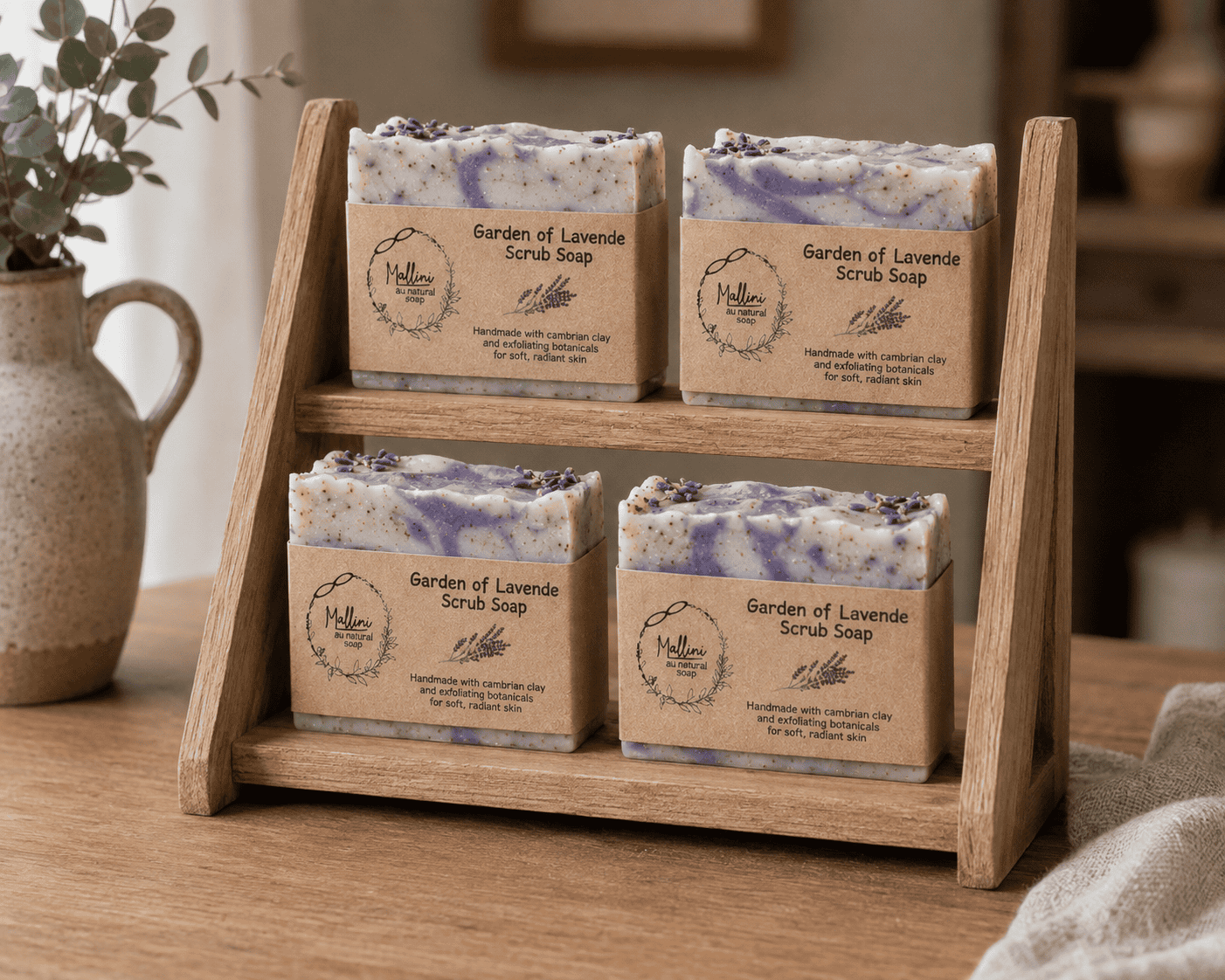 Four bars of lavender scrub soap in clear wrap, displayed on a two-tier wooden stand. Each bar has a kraft paper label reading 'Garden of Lavender Scrub Soap' with a circular logo.